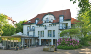 Hotelgebäude mit weißer Fassade, rotem Dach, verglastem Eingangsbereich und umliegender Gartenanlage mit blühendem Rhododendron.