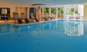 Caroline_Slider_Home_Pool Innenpool mit Liegestühlen, großen Fenstern und heller Decke in einem Wellnessbereich.