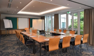 Konferenzraum mit U-förmig angeordneten Tischen, orangenen Stühlen, Leinwand und Tageslicht durch große Fenster.