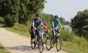 Celle_Radfahren_Familie_Aller_Angebotsbox Zwei Erwachsene und ein Kind fahren mit Fahrrädern auf einem Weg am Wasser.