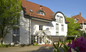 Hotelgebäude mit rotem Dach, geschwungenen Gauben und blühenden Sträuchern.