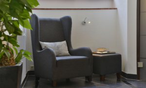 Leseecke mit grauem Ohrensessel, kleinem Tisch und Wandlampe neben Pflanze.