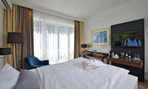 Zimmer mit Doppelbett, Fernseher, Schreibtisch und bodentiefem Fenster mit Vorhängen.