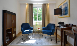 Sitzbereich mit zwei blauen Sesseln, Tisch mit Rose und Blick auf bodentiefe Fenster.