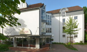 Hotelaußenansicht mit weißer Fassade, Glasfront und Eingangsbereich mit Bäumen.