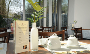 Frühstückstisch am Fenster mit Speisekarte, Teeservice und Rose in Vase.
