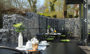 Hotel_Caroline_Mathilde_Celle_Fruehstueck_0005 Terrassentisch mit Kaffee, Tassen, Milch und Blick auf Steinmauer und Bäume.