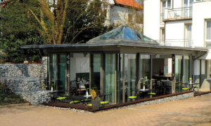 Hotel_Caroline_Mathilde_Celle_Fruehstueck_0410_073_ret Glaspavillon mit Terrasse, Sitzplätzen und umliegender Natur auf dem Hotelgelände.