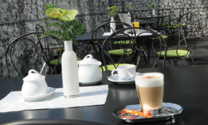 Hotel_Caroline_Mathilde_Celle_Fruehstueck_0410_087 Außentisch mit Kaffee, Milch, Rose in Vase und Stühlen vor einer Steinmauer.