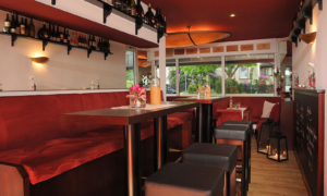 Hotel_Caroline_Mathilde_Celle_Restaurant_Bistro_Kanape_0030 Barbereich mit roten Hockern, Holztheke, Gläsern und Fensterfront.