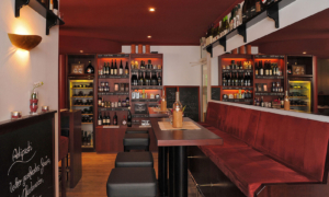 Hotel_Caroline_Mathilde_Celle_Restaurant_Bistro_Kanape_0031 Weinbar mit langen Holztischen, roten Polsterbänken, Barhockern und gefüllten Regalen.