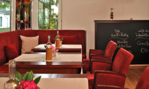Hotel_Caroline_Mathilde_Celle_Restaurant_Bistro_Kanape_0511_115 Gastraum mit roten Polsterbänken und -stühlen, gedeckten Holztischen, Flaschen und Tafel im Hintergrund.
