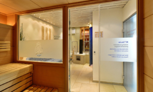 Hotel_Caroline_Mathilde_Celle_Sauna_Wellness_0023_2 Holzsauna mit Glaswand, Blick in den Ruhebereich und Logo auf der Tür.