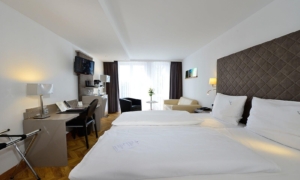 Zimmer mit Doppelbett, Schreibtisch, Fernseher, Sesselgruppe und Tageslicht durch bodentiefe Fenster.