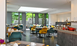 Großer, heller Frühstücksraum mit farbigen Stühlen, Buffet und Blick nach draußen.