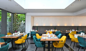 CM_Frühstück_0525_23 Frühstücksraum mit bunten Stühlen, gedeckten Tischen und Oberlicht.